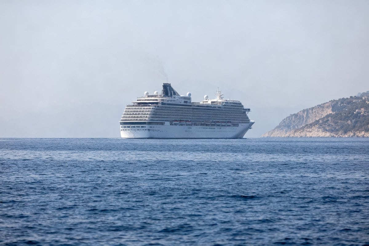 Un immense paquebot de croisière navigue au large d'une côte méditerranéenne sous un ciel d'été.