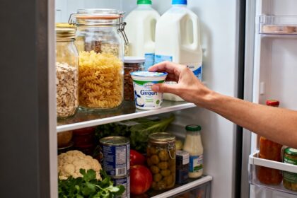Main saisissant un pot de yaourt dans un réfrigérateur garni de produits alimentaires du quotidien.