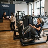 Femme sexagénaire sur une presse à cuisses dans une salle de sport, un homme au rameur et l'accueil en arrière-plan
