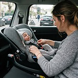 Une mère attache le harnais d'un bébé endormi dans un siège-coque gris dos à la route, fixé à l'arrière d'une voiture