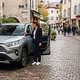 Toyota RAV4 gris garé dans une rue pavée, une femme debout près de la portière conducteur ouverte