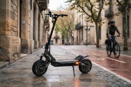 Trottinette électrique noire à grosses roues crantées, garée sur un trottoir mouillé le long d'une piste cyclable