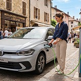 Femme branchant une Volkswagen grise sur une borne de recharge devant une boulangerie, dans une rue pavée