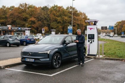 SUV électrique Volvo gris foncé branché à une borne Ionity sur une aire d'autoroute, un homme consulte son téléphone